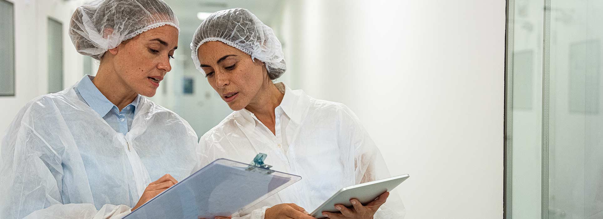 Two female colleagues in protective clothing studying a document in a light medical corridor