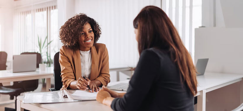 Two women sitting at a desk, one is smiling towards the other