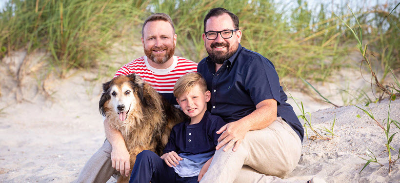 a family photo of Josh Stewart, his husband and son with their dog on beach dunes