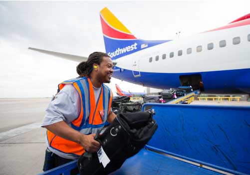 Image of a male ramp agent smiling while moving a bag from a baggage cart to a belt. He is in front of a Southwest Airlines airplane.