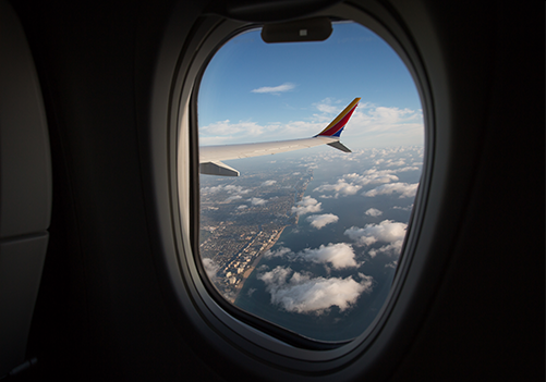 The view from a Southwest window seat. The Passenger sees a coast line, clouds, and a red, yellow, and blue Southwest wingtip.