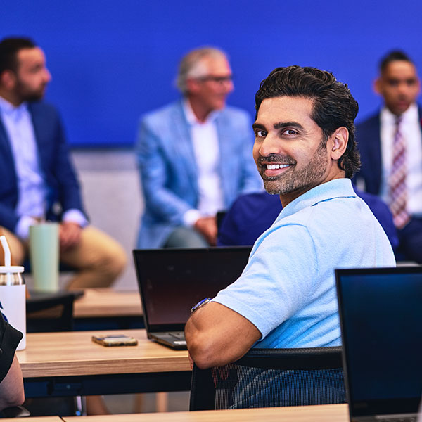 Student sitting at a large, turning around to smile.  Guest speakers in the background.