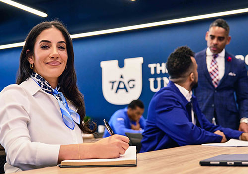 Students in a TAG U classroom, with a woman in the foreground holding a pen, smiling at the viewer