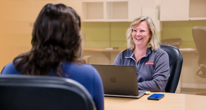 One employee smiles and talks to another while sitting at a desk.