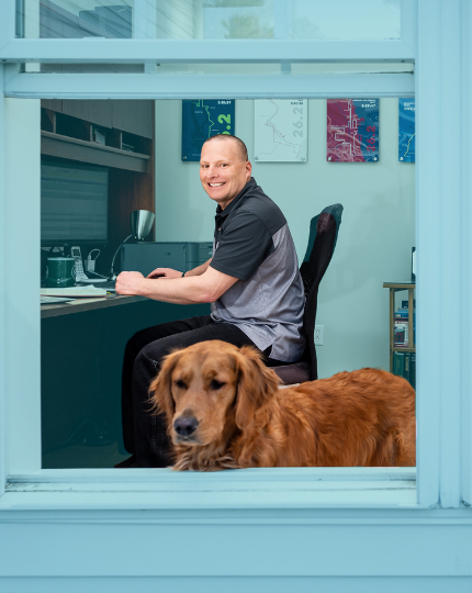 Greg, an engineering director, smiles for a photo in his home office. His golden retriever Luna, stands in the foreground.