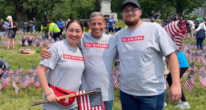 Three BAE Systems employees at a community investment event honoring our nations service members. The employee on the left is holding a few small American flags that will be planted in the ground.