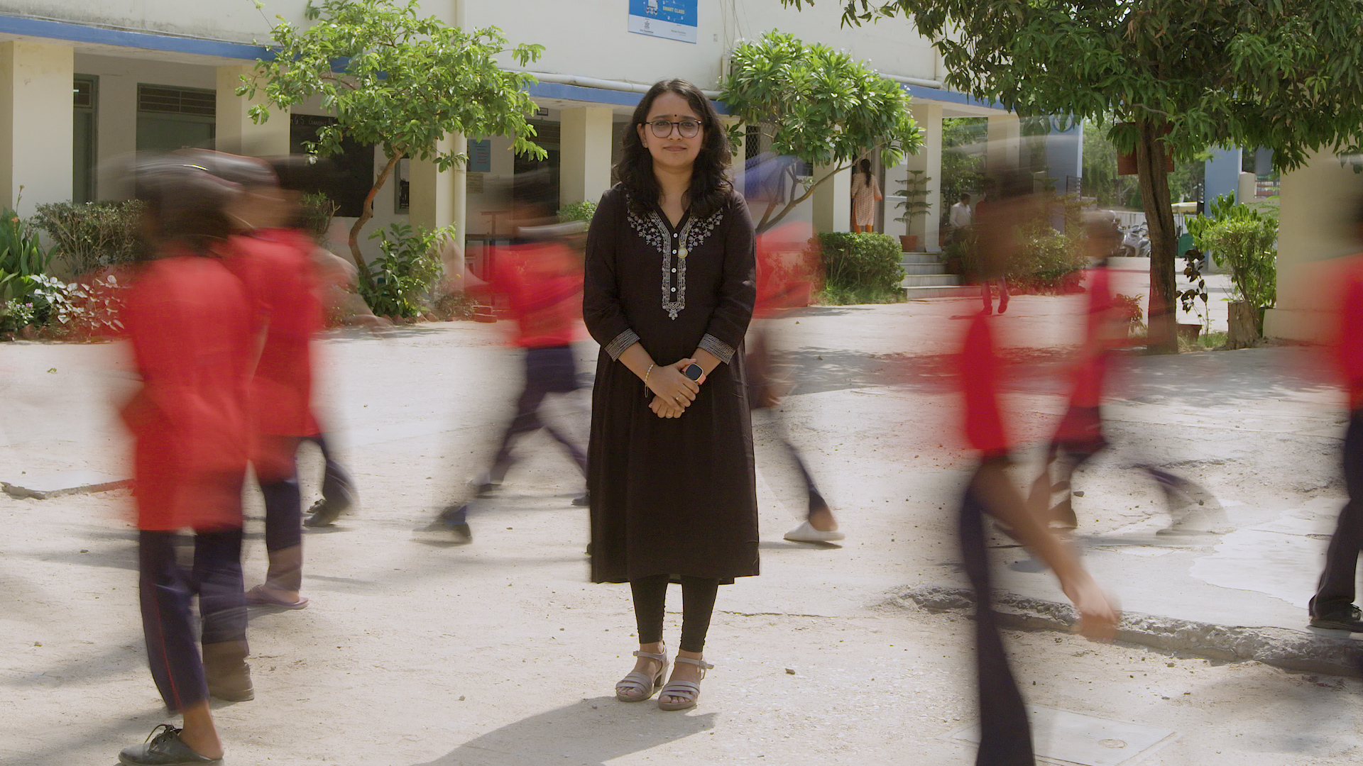 A woman stands still and centered in an outdoor setting, likely on a school campus. She is wearing glasses and a black, long-sleeved tunic with decorative embroidery at the neckline. The background shows blurred motion of children in red uniforms walking past her, creating a contrast between her stillness and their movement. 
