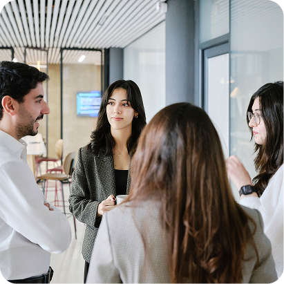 A group of four people in an office are situated in a circle and appear to be conversing with one another. There are three women and one man.