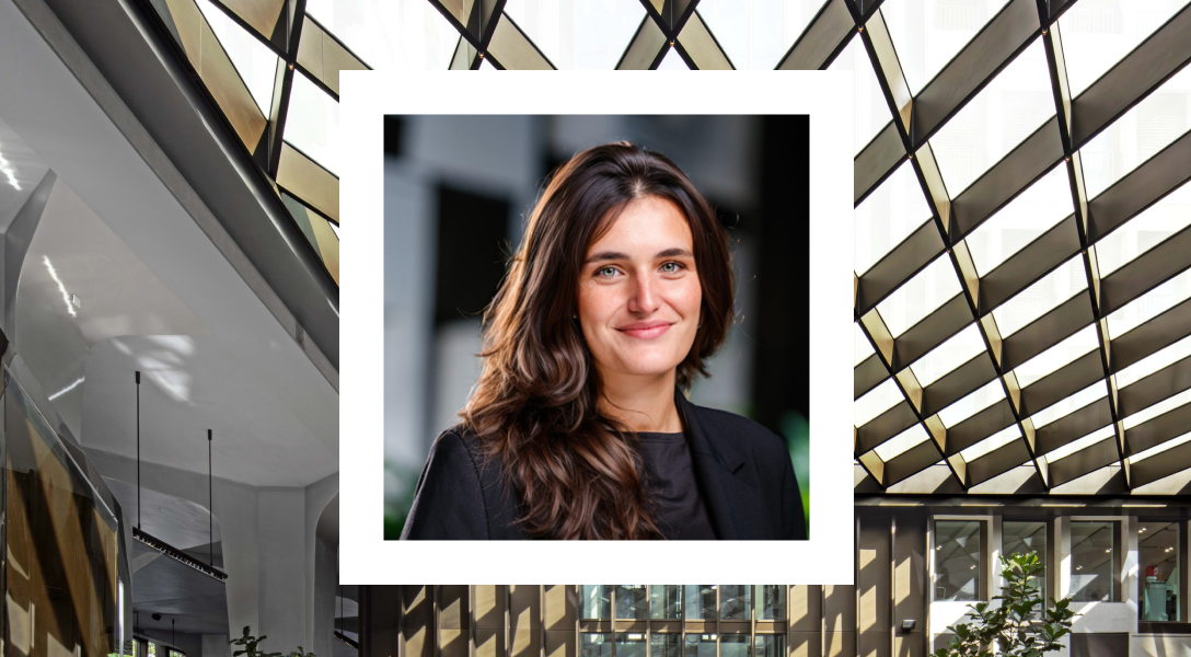 Portrait of a woman with long brown hair wearing a black blazer, smiling softly, set against a blurred background. Her photo is framed within a white border that contrasts with a modern architectural setting in the background, featuring large glass panels and angled beams, creating a geometric pattern with natural light streaming in.