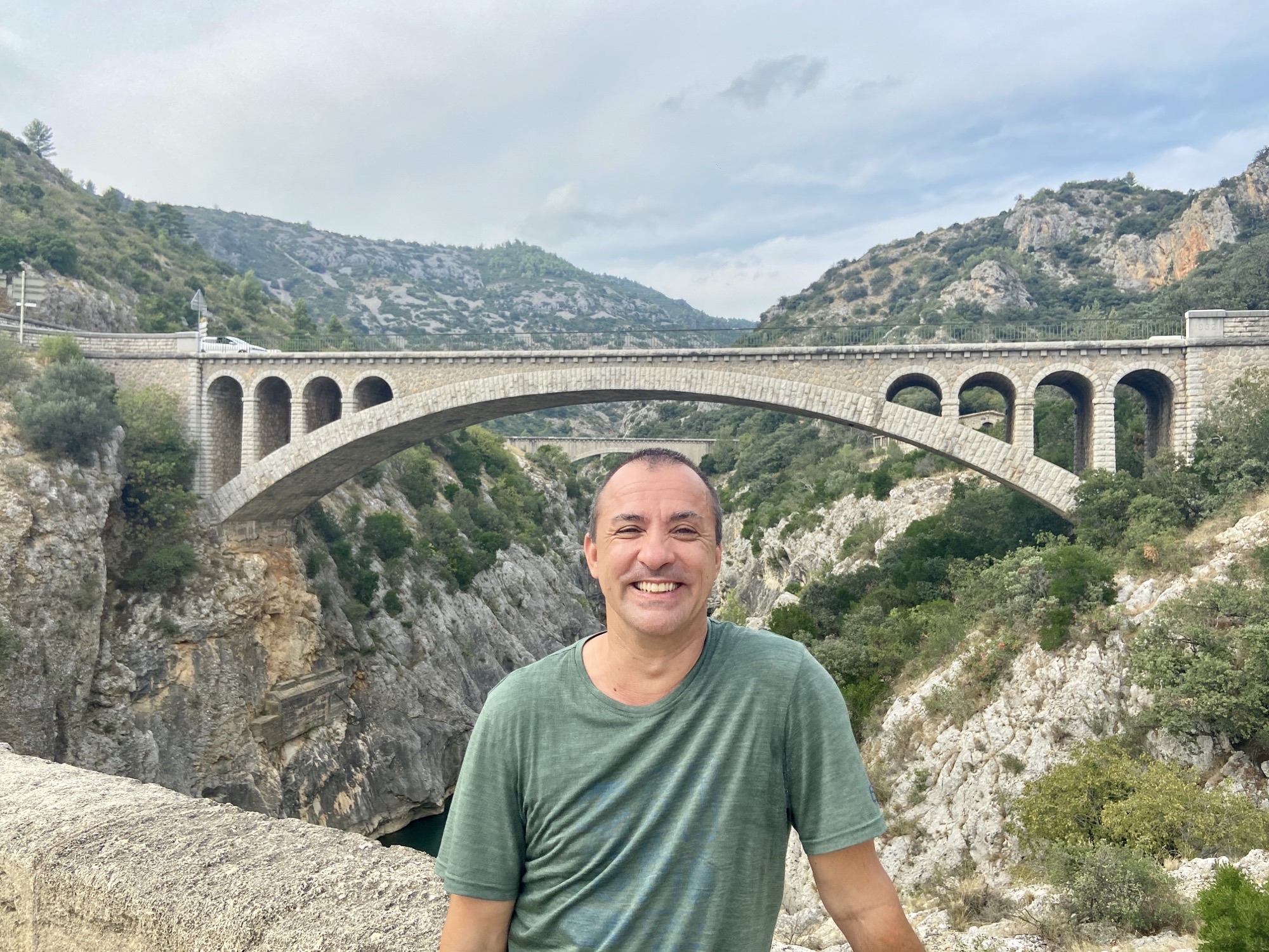 A man wearing a green t-shirt is standing and smiling in front of a stone bridge that spans across a deep gorge. The background features rocky cliffs covered with greenery and a cloudy sky. The bridge has multiple arches and extends across the entire width of the image, with mountains visible in the distance.