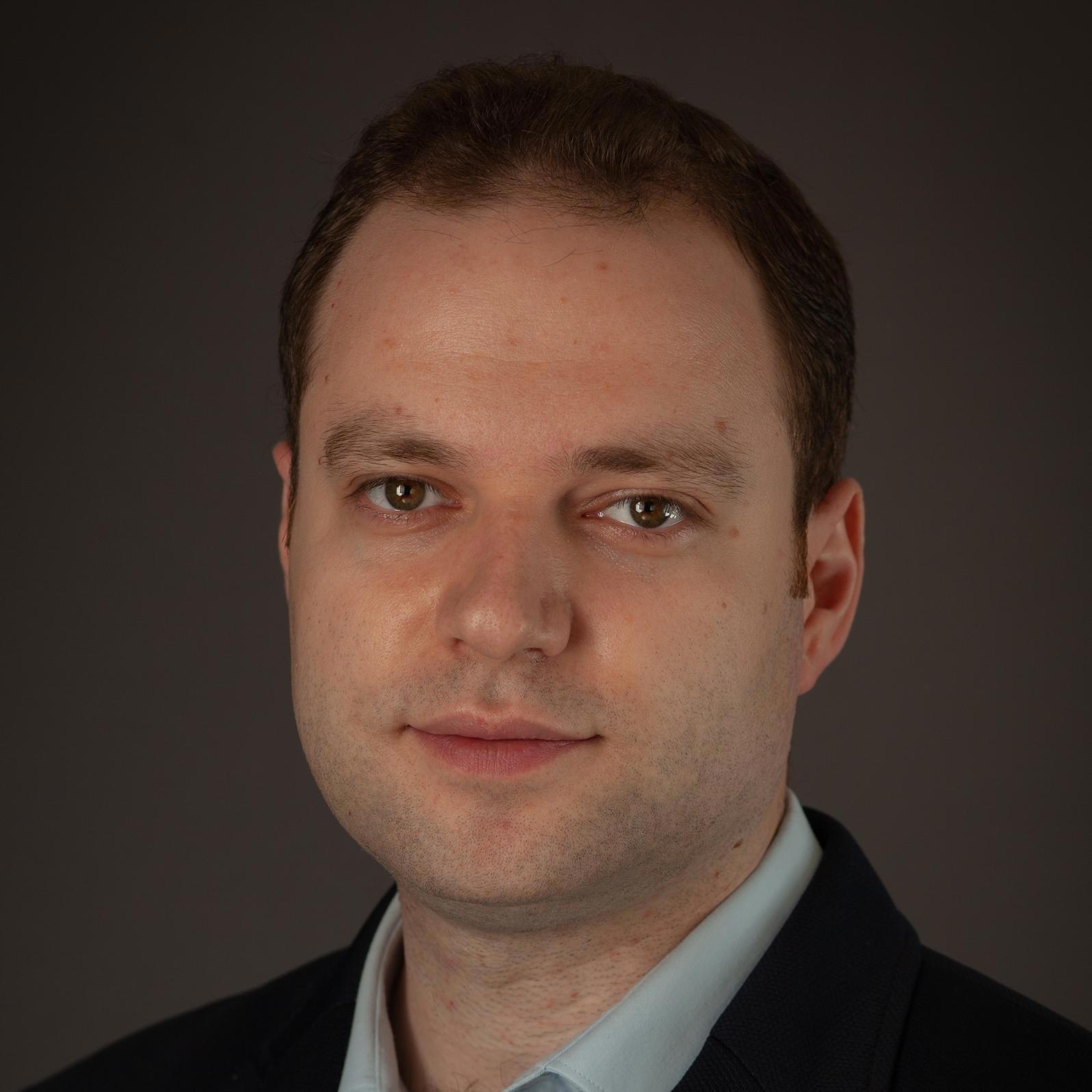 Gros plan d'un homme aux cheveux courts châtain foncé et à la peau claire. Son expression est neutre et il regarde droit vers l'objectif. Il porte une veste de costume sombre sur une chemise à col bleu clair. L'arrière-plan, sombre et uni, met son visage en valeur.