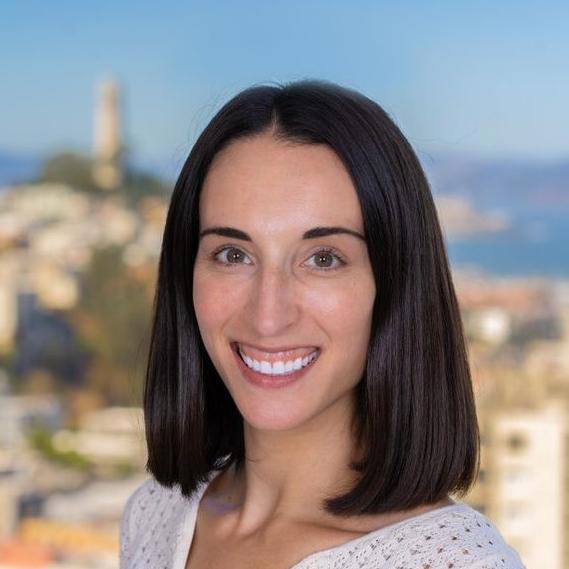 A smiling woman with straight, shoulder-length dark hair stands against a backdrop of a scenic cityscape with blurred buildings and a distant view of the water. She is wearing a white top and her friendly expression and bright smile exude warmth and approachability. The sunny background adds a touch of vibrancy to the portrait.