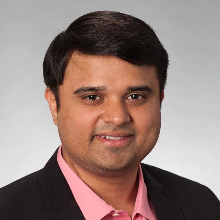 A smiling man with short, dark hair wearing a pink collared shirt and a dark blazer stands against a plain gray background. His friendly and professional demeanor is highlighted by his confident expression, creating a welcoming and approachable impression.