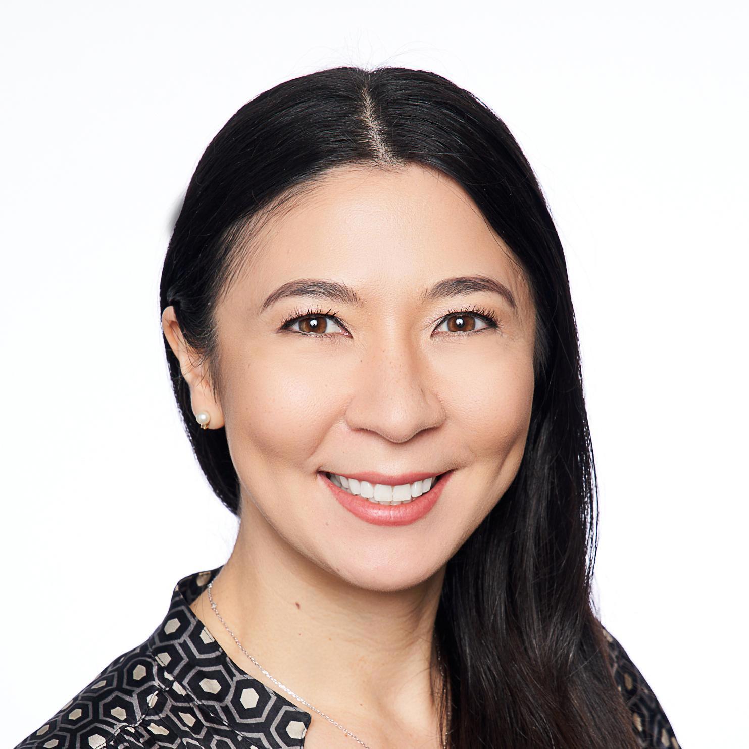 A smiling woman with long, dark hair wearing a patterned blouse stands against a plain white background. Her friendly and professional demeanor is highlighted by her bright eyes and warm smile, creating a welcoming and approachable impression.