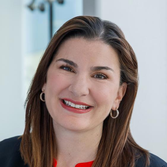 A smiling woman with shoulder-length brown hair, wearing hoop earrings and a red top with a dark blazer, stands indoors with a soft background. Her warm and approachable demeanor is highlighted by her bright smile and welcoming expression.