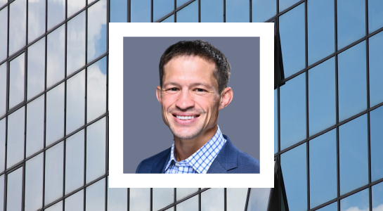 A smiling man wearing a blue suit jacket and a light blue checkered shirt. He has short dark hair and is set against a plain gray background. The image is framed within a white border and overlaid on a background featuring the reflective glass facade of a modern building with a partly cloudy sky visible in the reflection.