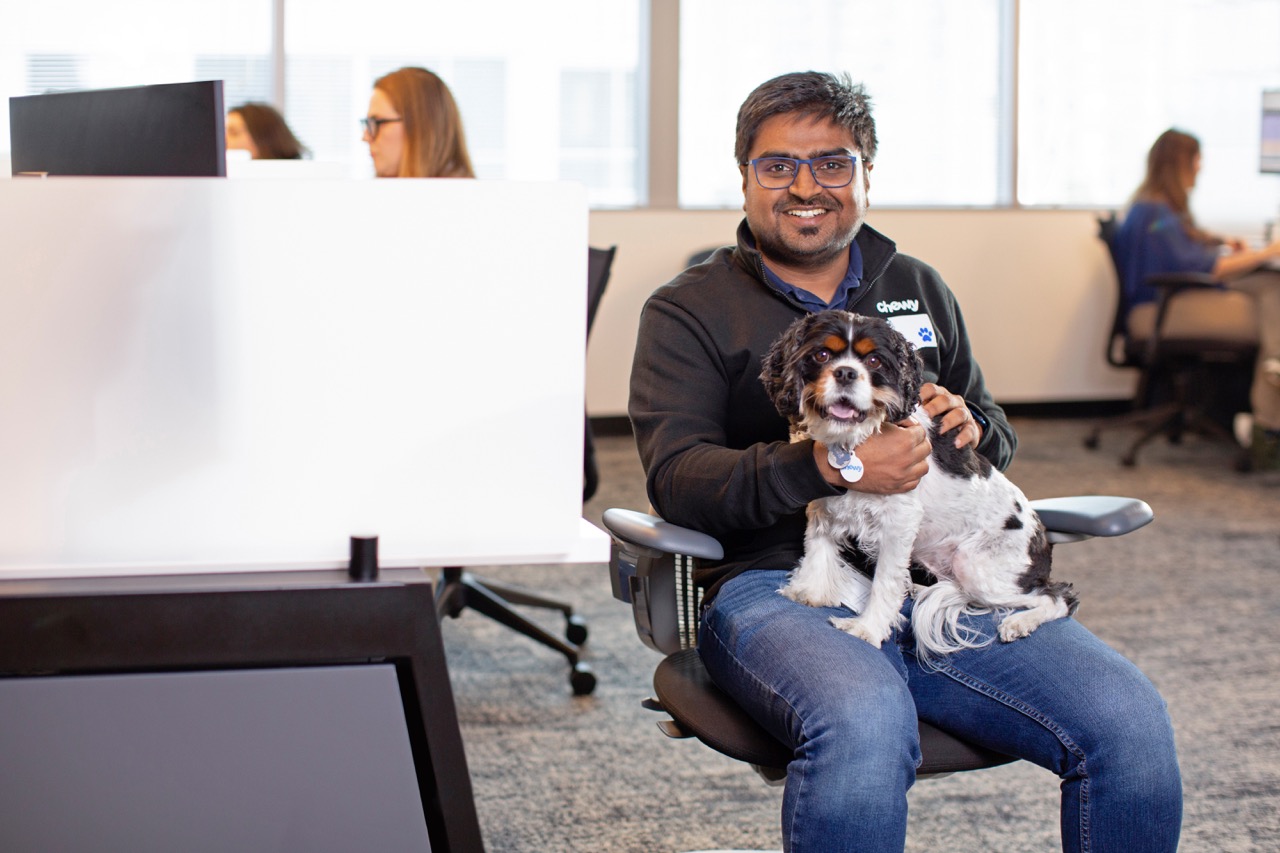 A black and curly dog giving its paw in exchange for a treat from a Chewy team member sitting in a chair
