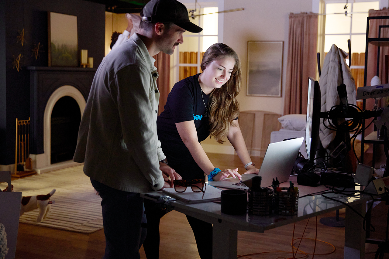 A smiling Chewy team member looking out the office window while another team member decorates a Christmas tree in the background