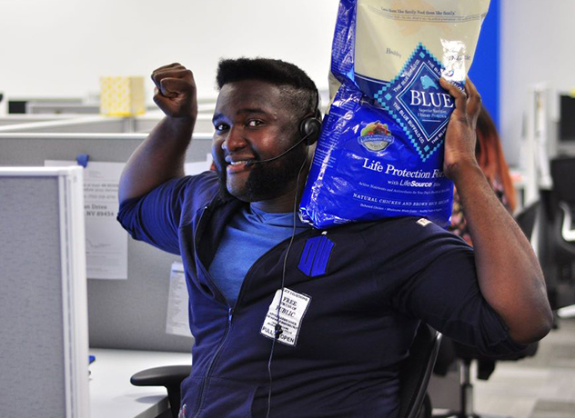 A Chewy team member smiling and flexing their muscles while holding a bag of dog food on their shoulder