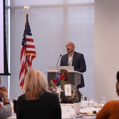 Children’s employee and veteran stands in front of United States flag during Veterans breakfast.