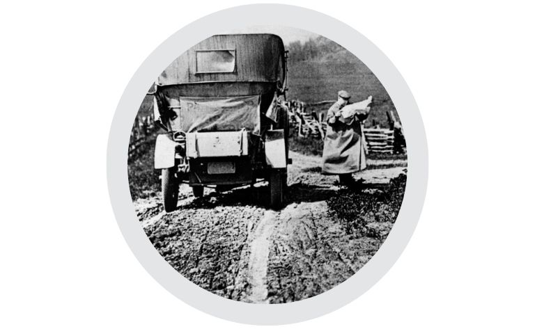 Historic black‑and‑white photograph showing an early automobile on a muddy rural road with a person standing nearby