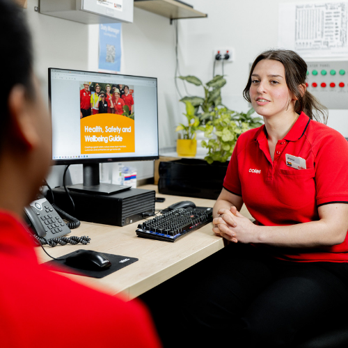 Two supermarket team members speaking in the office, a computer in the background shows a health and wellbeing learning page. 