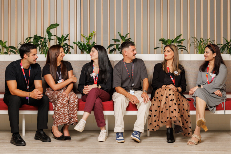 A diverse group of team members sitting together on a modern red bench in a bright, welcoming office space with green plants in the background. They are wearing workplace lanyards and engaging in conversation, reflecting collaboration and inclusion.