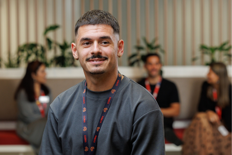 A team member wearing a company lanyard seated in a modern office lounge area with plants and wooden paneling in the background. Other colleagues are visible in conversation behind, creating a collaborative and welcoming environment.
