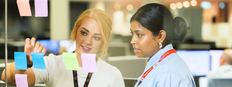 Two team members collaborating in an office space, reviewing colourful sticky notes on a glass wall. The scene reflects brainstorming and teamwork in an open-plan environment.