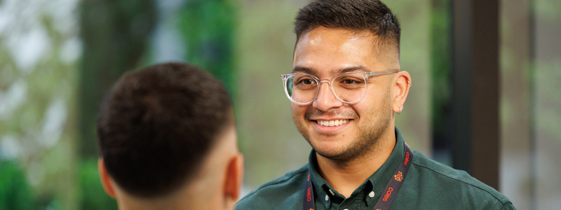 Two colleagues having a conversation in a bright, modern workspace with greenery visible in the background. Both are wearing company lanyards, highlighting a professional and approachable setting.