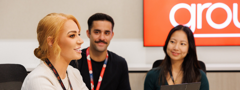 A group of colleagues sitting together in a modern meeting room, wearing company lanyards and engaging in discussion. A bright red sign with partial text is visible in the background, creating a vibrant and collaborative atmosphere.