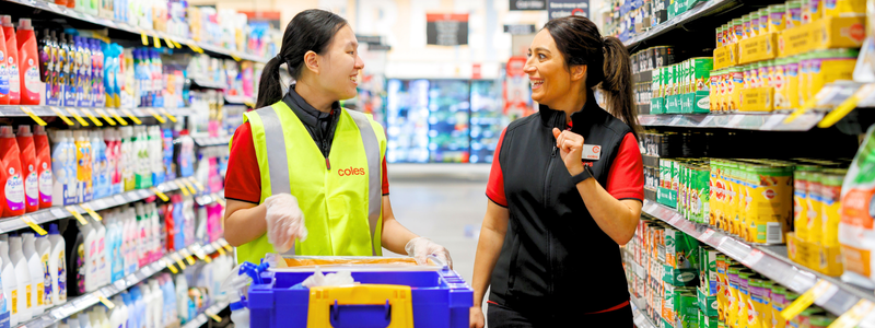 Two female store team members working in a supermarket aisle. The female team member to the left wears a high vis vest and pushes a cleaning trolley. The female team member to the right wears a black branded Coles vest.
