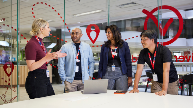 A variety of corporate team members gathered in a meeting room. They stand together around a table and laptop talking and smiling.