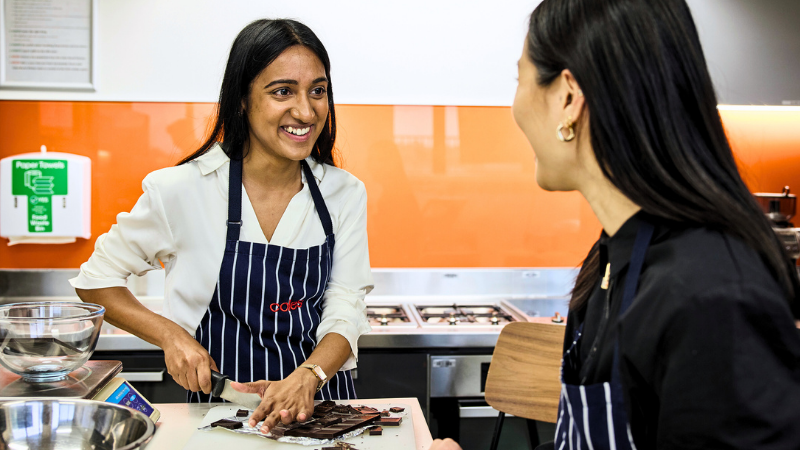 Two Commercial and Merchandise Coles team members working together in Coles SSC kitchen. Both team members are wearing Coles branded aprons