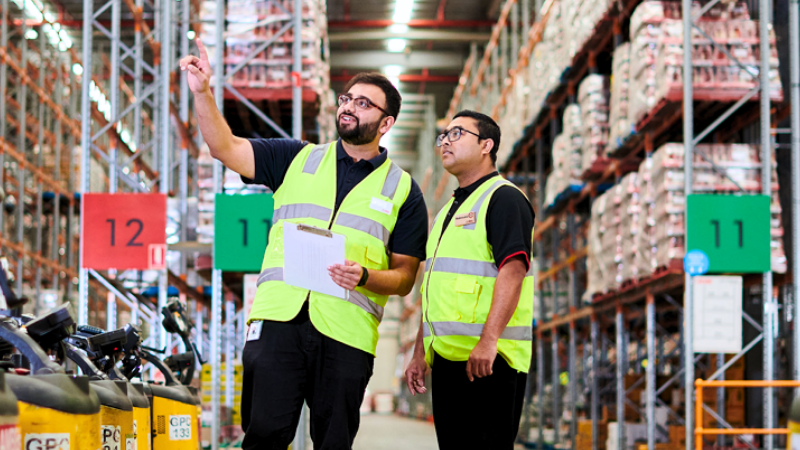 Two males wearing Coles branded high vis vests within a Coles warehouse. 