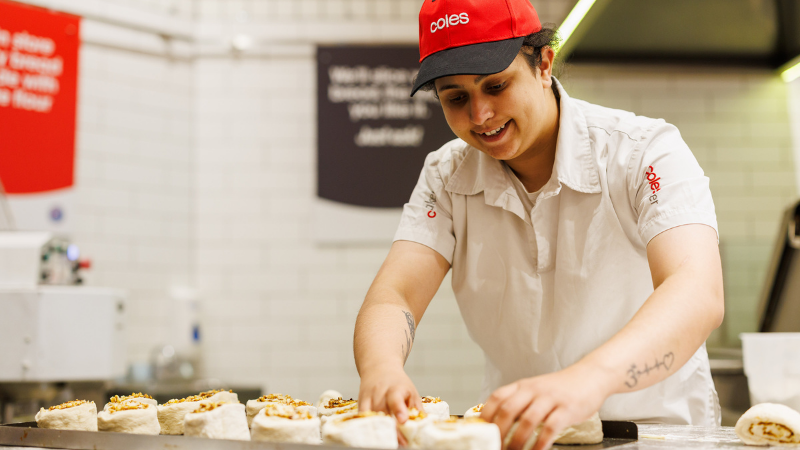 A female Coles Baker is seen preparing dough.  She is wearing a Coles branded red cap and white uniform
