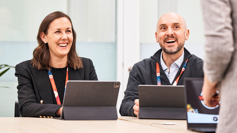 A female and male team member sitting side by side laughing in a meeting