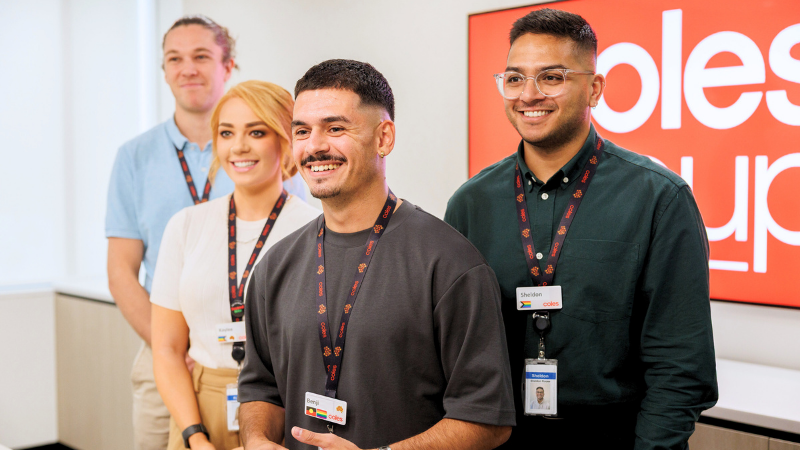 Four Coles graduates standing together smiling in front of a Coles Group sign