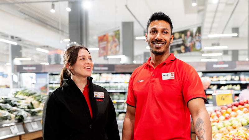 Two team members standing in fresh produce section of a Coles Supermarket. One team member wears a black branded Coles jacket while the other wears a Coles branded red polo.