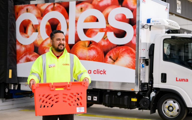 A Coles team member in a high-visibility jacket holding a red delivery crate in front of a branded delivery truck, showcasing commitment to safe and efficient online order fulfilment.