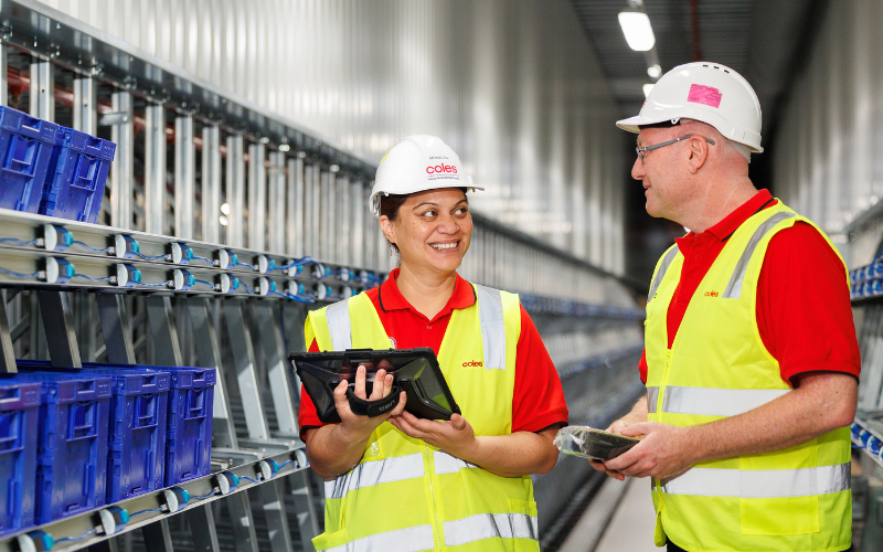 Two Coles team members wearing safety vests and helmets in a distribution centre, using digital devices to manage operations, highlighting innovation and workplace safety.