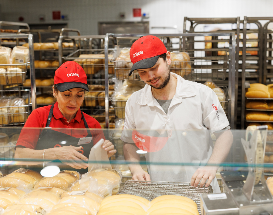 Two Coles bakers working together in the bakery. The Female baker wears a red polo, a black Coles-branded apron, and a red cap. The male baker wears a white Coles-branded uniform and a red cap. Both are sorting the loafs of bread behind a glass counter.