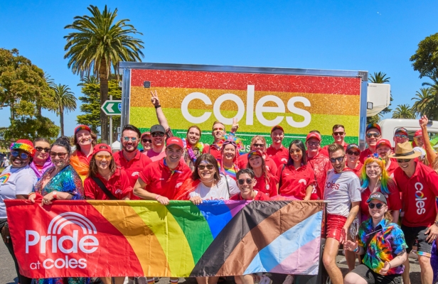 Coles Team members celebrating pride outside in the sunshine holding a  large Pride at Coles flag in front of a Coles van  that has been decorated with rainbow colors