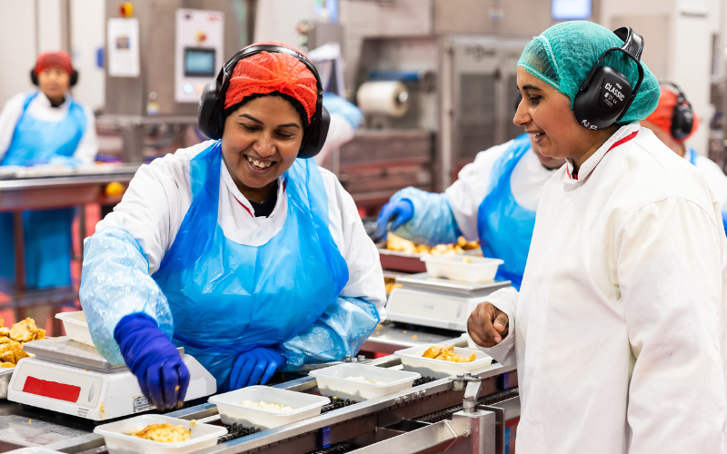 Two female team members preparing meals on a conveyor belt machine.  Each has safety headphones on, blue plastic vests and white coats and hair nets on their heads