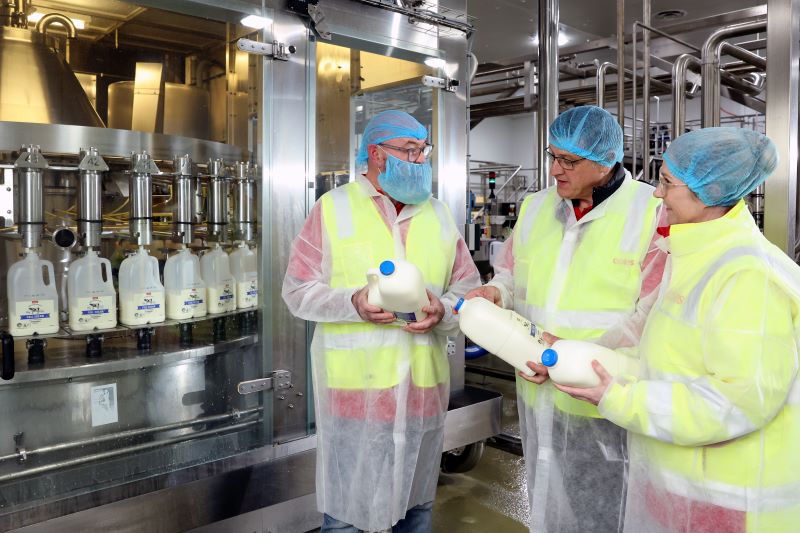 Three team members inside a fresh milk processing facility, with machinery in the background dispensing milk into plastic bottles