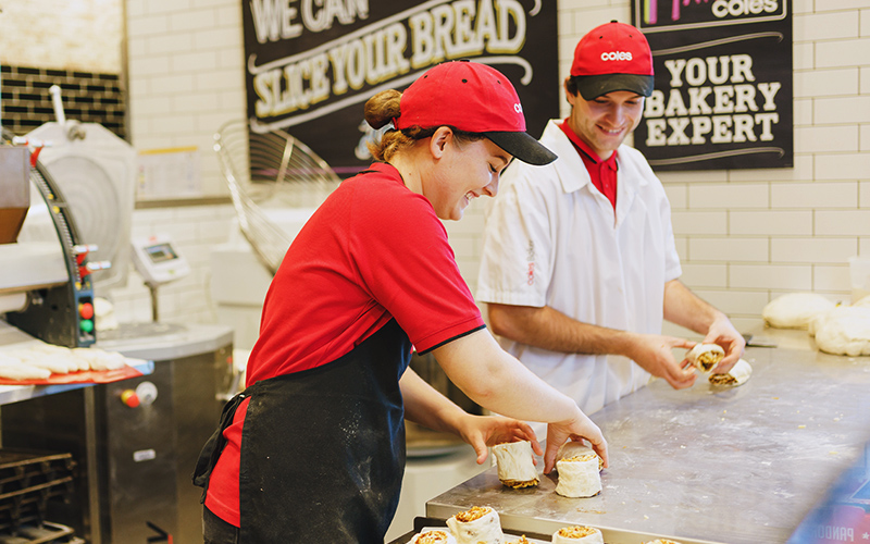 Two Coles team members working together in the bakery section, preparing fresh bread rolls on a stainless steel counter. Both are wearing red Coles caps and branded uniforms, with bakery equipment and signage in the background promoting fresh bread and expert service. The image highlights collaboration, skill, and commitment to quality in an inclusive workplace.