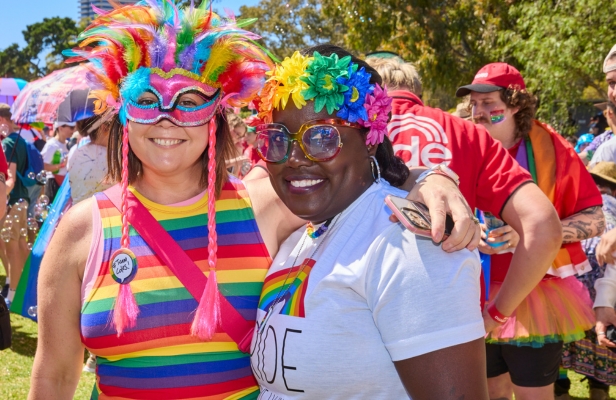 Two women wearing colorful feathered headdresses and embracing each other outdoors at a Pride festival, in bright sunshine with people celebrating in the background