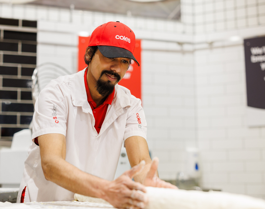 A Male baker wears a white Coles-branded uniform, and a red cap is seen kneading dough inside a Coles Bakery