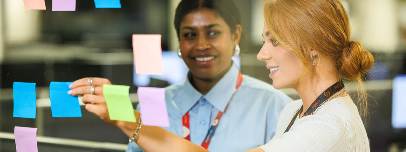 Two female corporate team members sticking post-it notes to the window within the Store Support Centre
