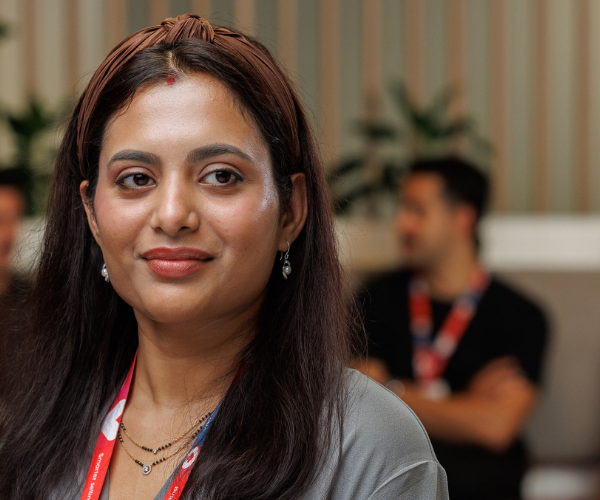 A team member wearing a company lanyard and layered necklaces, standing in a modern office lounge with plants and wooden paneling in the background.